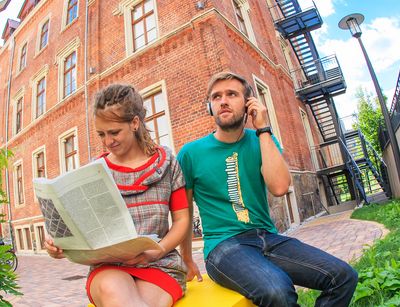 Eine Studentin und ein Student sitzen vor einem Studentenwohnheim, einem historischen Gebäude mit Ziegelfassade. Sie liest Zeitung, er trägt Kopfhörer.Foto: Studentenwerk Leipzig