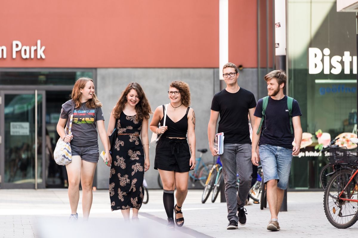 zur Vergrößerungsansicht des Bildes: Studierende gehen nebeneinander auf dem Campusgelände