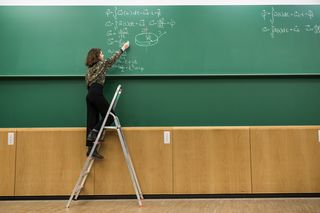 Studentin steht auf einer Leiter und schreibt Formeln an die Tafel eines Hörsaals, Foto: Christian Hüller