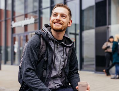 Studienbotschafter Michael sitzt auf einer Bank auf dem Campus und trinkt Kaffee, Foto: Christian Hüller