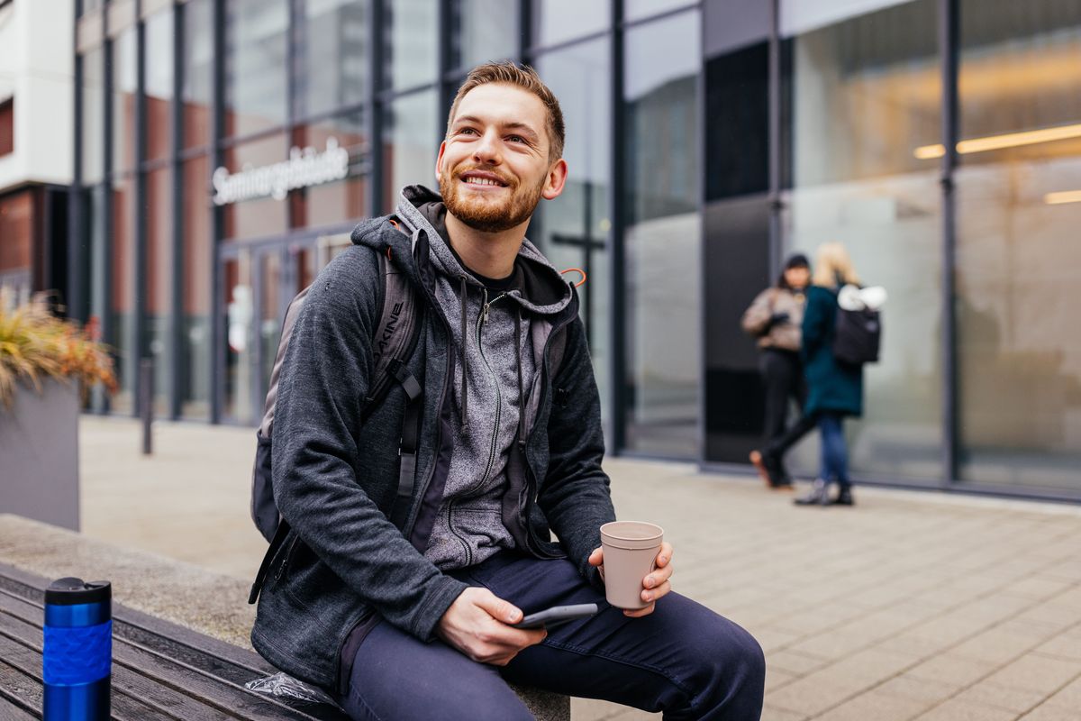 zur Vergrößerungsansicht des Bildes: Studienbotschafter Michael sitzt auf einer Bank auf dem Campus und trinkt Kaffee, Foto: Christian Hüller