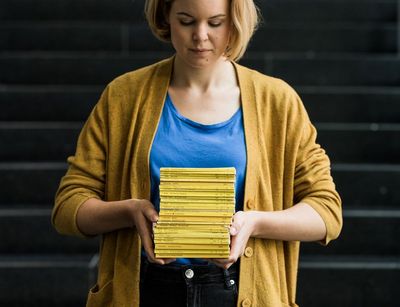 Studentin hält gelben Bücherstapel vor sich und blickt darauf, Foto: Christian Hüller