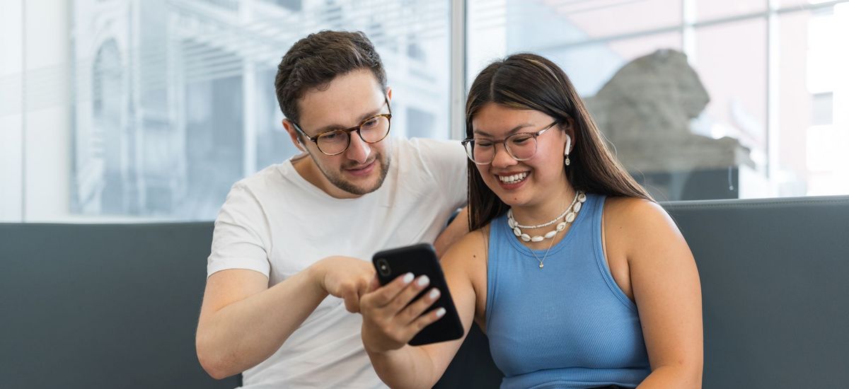 Ein Student und eine Studentin sitzen eng beieinander, schauen gemeinsam auf ein Handy und haben Earbuds in den Ohren, Foto: Christian Hüller