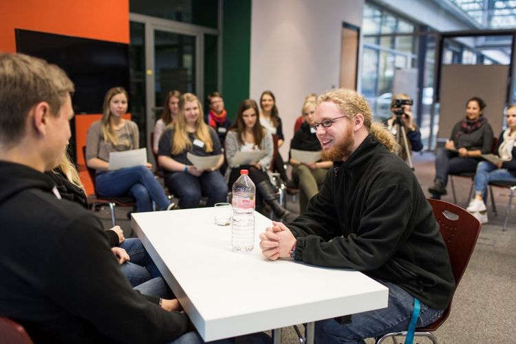 Studierende üben ein Gespräch. Zwei Studierende sitzen einem Studenten gegenüber, während die anderen Kommilitonen sich Notizen dazu mache, Foto: Christian Hüller