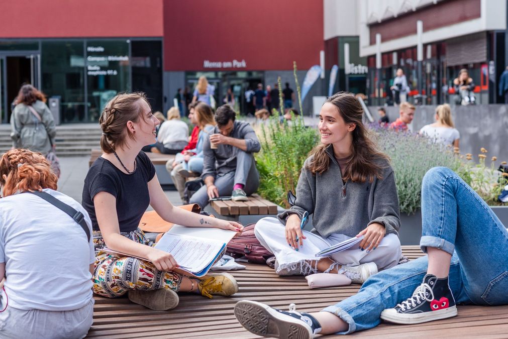 zur Vergrößerungsansicht des Bildes: Zwei Studentinnen sitzen auf einer Bank im Leibnizforum und sprechen miteinander, um sie herum herrscht reges Treiben im Uni-Innenhof, Foto: Kirsten Nijhof
