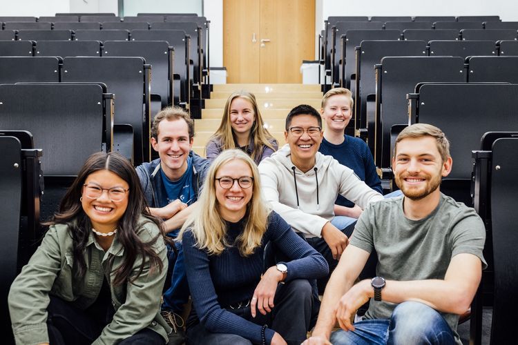 Farbfotografie von sieben Studierenden, den Studienbotschafterinnen, die im Gang, zwischen den Stuhlreihen eines Hörsaals auf dem Boden sitzen