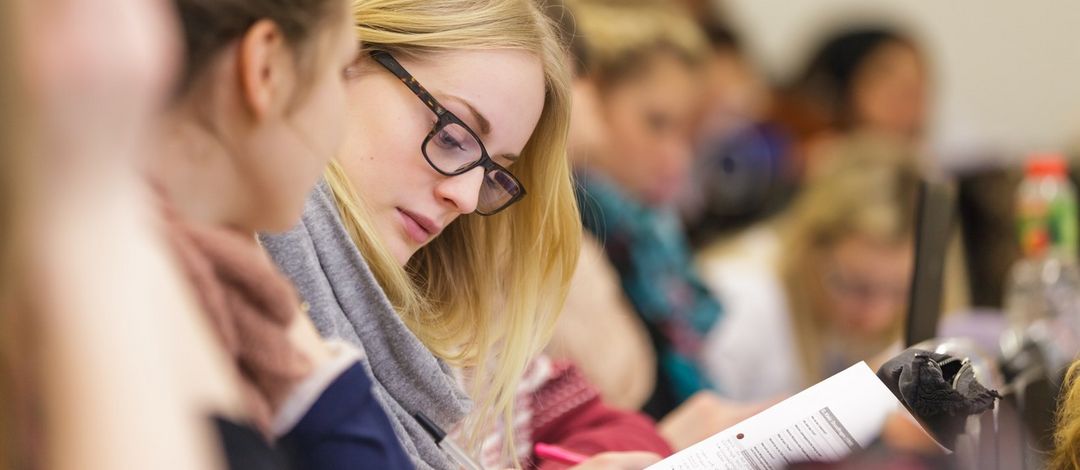 Im Blick ist eine Sitzreihe von der Seite mit Fokus auf eine Studentin, die gerade etwas aufschreibt, Foto: Christian Hüller