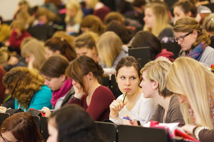 Blick auf die Sitzreihen in einem vollen Hörsaal und zwei Studentinnen reden miteinander, Foto: Christian Hüller