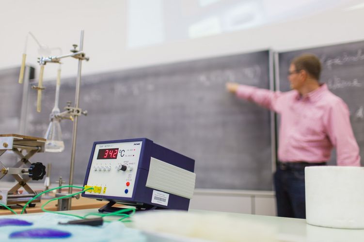 Blick auf ein Experiment mit unterschiedlichen Geräten. Im Hintergrund zeigt der Dozent auf die vollgeschriebene Tafel, Foto: Christian Hüller