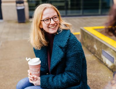 Studienbotschafterin Patricia sitzt auf einer Bank auf dem Campus und hält einen Becher in der Hand, Foto: Christian Hüller
