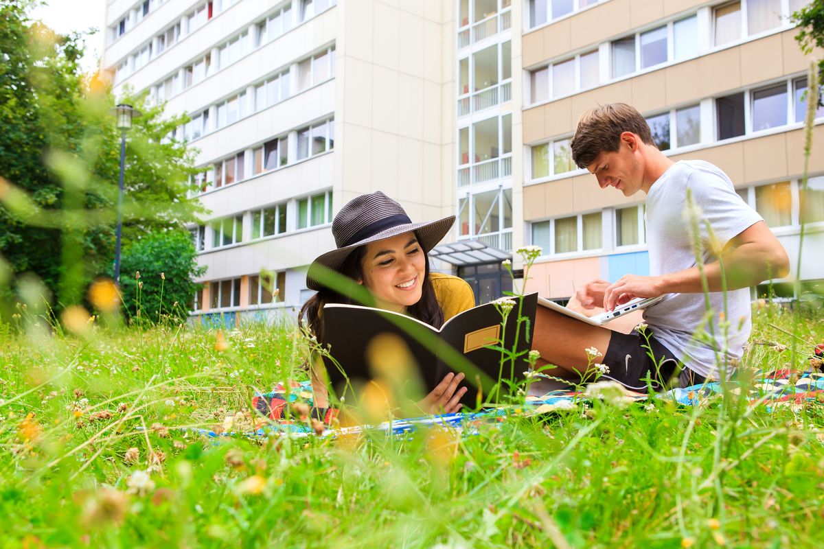 zur Vergrößerungsansicht des Bildes: Zwei Studierende sitzen auf der Wiese vor einem Studentenwohnheim.