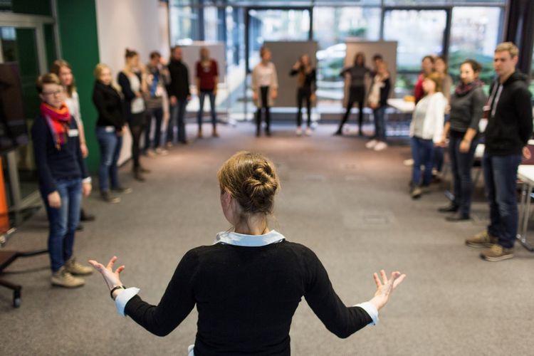 Fokus auf eine Frau, die mit dem Rücken zur Kamera steht und zu Studierenden spricht, die vor ihr im Kreis stehen, Foto: Christian Hüller