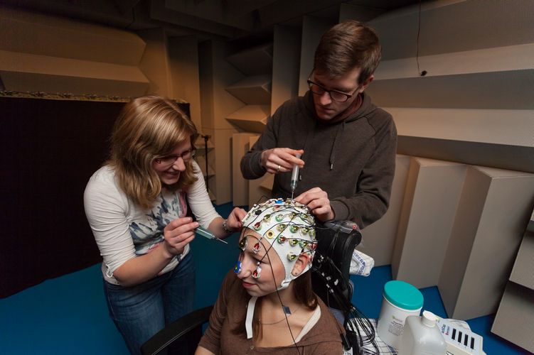 Zwei Studierende sind in einem schallisolierten Raum und führen ein Experiment an einer Probantin durch, die eine Kopfhaube mit Kabeln trägt, Foto: Christian Hüller