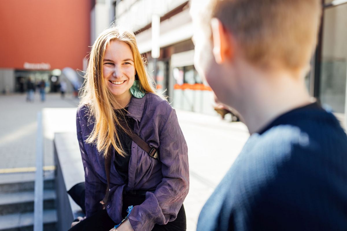 zur Vergrößerungsansicht des Bildes: Studienbotschafterin Natalie lächelt beim Gespräch auf dem Campus, die Sonne scheint im Hintergrund, Foto: Christian Hüller